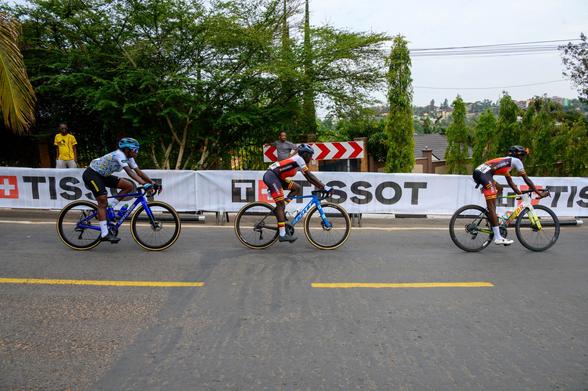 A group of cyclists in competitive attire are riding in a race, seen from behind. They are navigating a curved road, with multiple bicycles in motion showing various colors and styles. The setting appears outdoors with trees in the background.