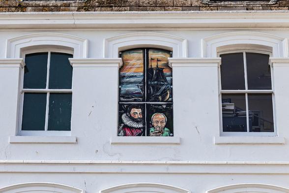 We're looking at an upper floor of a building, painted white but in need of a touch up to repair some flaking and cracked areas. A thin strip of tan-coloured roof tiles just frames the top of the shot but we're mainly focussed on the three windows arranged horizontally across the centre of view. Each window is broadly identical, being a tall rectangular frame in which there are four evenly-sized window panes. The windows appear to be sash-opening but all are closed and the upper edge has a slight convex curve to it, not enough to quite count as an arch. Relief rail work around the frames and between each window gives the impression that each one is just behind tall crenellations of some fortified house. The left window is bare, each pane just black. Within the darkness of the room behind the right window we can just make out office equipment and the edge of a door on the far wall. The middle window is filled with painted artwork. The scene is of a sunset sky of orange over the dark blue sea and the silhouette of a tall-masted sailing ship. Two characters are at the bottom as if looking out on the street below. The bearded man on the left wears Elizabethan clothing in red with a large white ruff and is clearly Sir Francis Drake. The older man on the right seems more modern, wearing a plain green top and drinking from a large brandy glass.