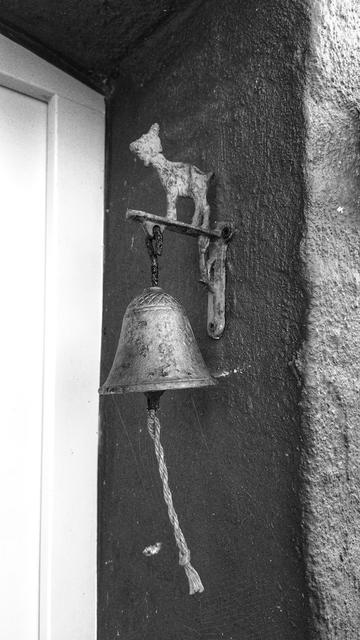 Eine kleine Haustürglocke, die etwa auf Kopfhöhe außen vor der Haustür an der Mauerleibung angebracht ist. Auf der Metallkonsole, an der die Glocke hängt, steht eine kleine Tierfigur aus Metall in Umrissen, vermutlich ein Lamm. Unten aus der Glocke hängt ein kurzes Stück Kordel, das offensichtlich leicht im Wind schwingt. Das Bild ist in schwarz-weiß.