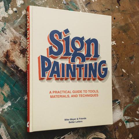 Photo of a hardback book on a paint-spattered surface. The cream cover of the book is dominated by the hand-lettered title “Sign Painting”.