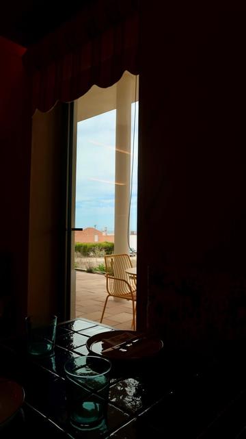 A view from a dimly lit room, with two blue glasses and a plate visible in the foreground, looking through an open doorway onto a sunny, tiled patio with a yellow wire chair and a glimpse of red tile roofs and the sky in the distance.