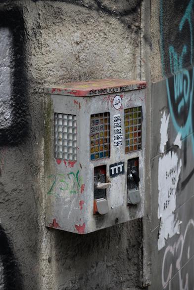 Ein verwitterter Kaugummiautomat mit zwei Fächern. Die rote Farbe ist kaum noch vorhanden, der Rest ist silbern. Oben steht in schwarz: "FÜR IMMER KIND BLEIBEN". Auf der Front noch ein paar Aufkleber zu sehen,  die Sichtfenster sind vergittert. Der Automat hängt an einer Hauswand, mit einigen Graffiti.