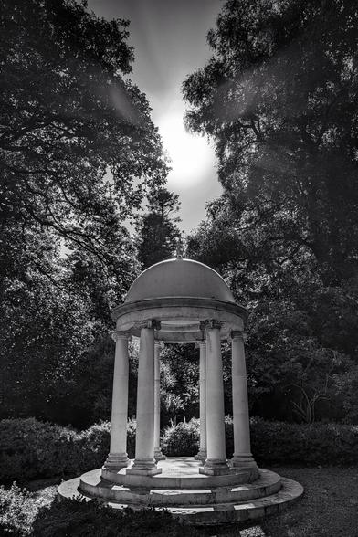 A striking black and white photo of a circular, classical-style stone temple or gazebo, featuring a domed roof supported by six columns. The structure is set on a low, tiered base in a garden or wooded area. Tall, dense trees frame the structure on both sides, with the bright sun directly behind the dome at the top centre, creating a strong lens flare and dramatic contrast.