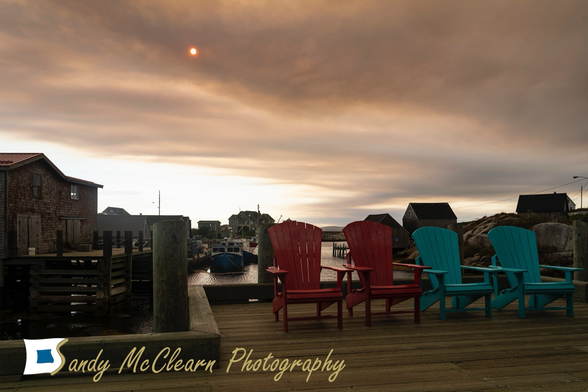 Sun made red by wildfire smoke over chairs on a wharf in a fishing harbour.