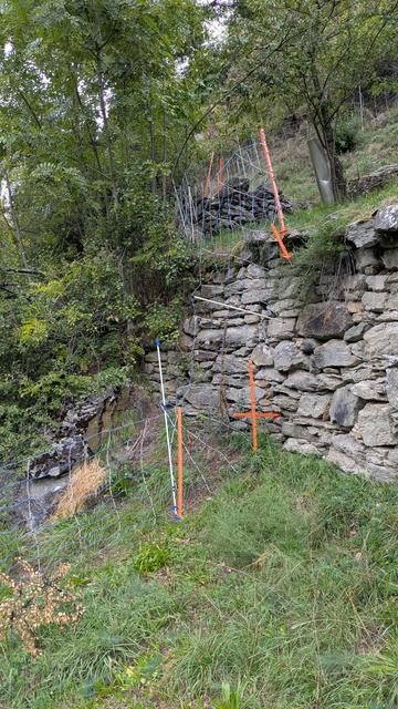 Ein Schafzaun (Flexinetz) führt direkt über eine 2m hohe Bruchsteinmauer. Direkt an die Mauer ran, umgefaltet, senkrecht die Mauer hoch und oben dann gerade weiter. Zusätzlich zu den Pfählen des Netzes halten organgefarbene Viehüterstecken das Netz auf Spannung und verhindern Erdschlüsse.
Der unterste Draht unserer Netze ist ein Erdungsdraht, deswegen darf der unterste Draht auf keinen Fall die anderen Drähte berühren.