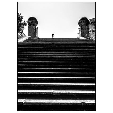 his black-and-white photograph captures a wide staircase leading upward, flanked by two large, dome-shaped structures at the top. A single person is seen walking down the stairs, positioned centrally in the frame. The staircase is symmetrical, with the steps creating strong horizontal lines that draw the eye toward the person and the domes. The sky above is clear, providing a stark contrast to the architectural elements. (draft by mistral.ai, edited by author)