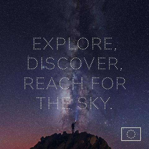 A wide-shot image of a starry night sky, with the Milky Way visible. A silhouetted person stands on a rocky peak, shining a flashlight upward. The text "EXPLORE. DISCOVER. REACH FOR THE SKY." is written in white dots across the sky. The EU emblem appears in the bottom-right corner.