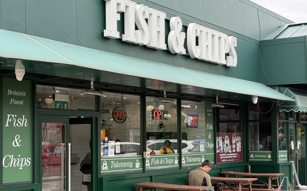 The front window of a fish and chip shop takeaway. A large sign above with a green background.