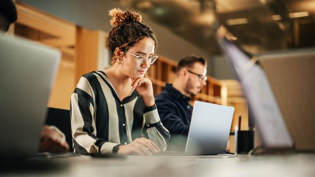 vrouw zit aan tafel achter laptop, tussen mensen die ook een laptop zitten