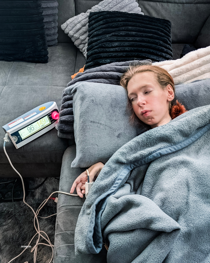 Ich liege erschöpft auf einem grauen Sofa, eingehüllt in eine weiche graue Decke. Neben mir steht ein medizinisches Gerät, das Sauerstoffsättigung und Puls anzeigt. Ein Sensor ist an meinem Finger befestigt. Um mich herum sind sehr viele Kissen.