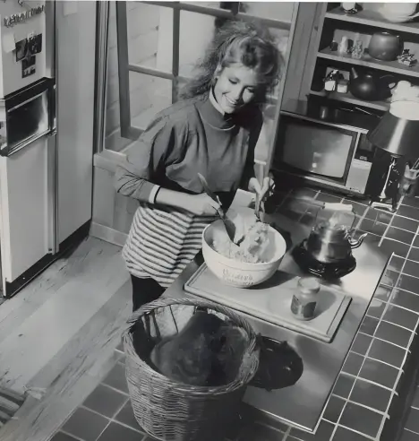 A black-and-white photograph from the mid-1980s showing Deidre Hall, best known as Marlena Evans on Days of Our Lives, standing in a domestic kitchen. She wears a dark, long-sleeved high-neck sweater, dark trousers, and a striped apron. Her curly, shoulder-length light brown hair is partially tied back. Deidre is mixing ingredients in a large bowl on the kitchen island and smiling warmly at a cat sitting in a basket on the counter. The kitchen features tiled countertops, wooden flooring, and various utensils and appliances. The image captures a cozy, candid moment that contrasts with the dramatic, high-stakes storylines of her soap opera character.