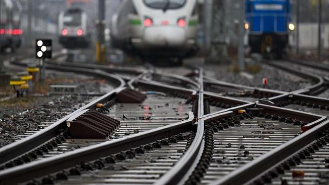 Das Bild zeigt eine Ansicht von Eisenbahnstrecken mit mehreren Gleisen, die sich kreuzen. Im Vordergrund sind die Gleise und die Schienen mit Steinen und Schwellen zu sehen. Im Hintergrund sind mehrere Züge zu erkennen, darunter ein moderner Hochgeschwindigkeitszug mit einer markanten, aerodynamischen Form und ein blauer Güterzug. Die Strecken sind in einem Industriegebiet, und es gibt Signalanlagen und andere Infrastrukturelemente. Die Umgebung wirkt nass, was auf Regen hindeutet. Die Szene vermittelt ein Gefühl von Aktivität und Komplexität im Eisenbahnverkehr.