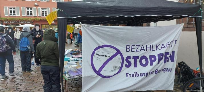 Foto von einem Infostand unter einem schwarzen Pavillon. Vorne hängt ein großes weißes Transparent auf dem in lila eine durchgestrichene Bezahlkarte in einem Kreis abgebildet ist, daneben steht "Bezahlkarten stoppen Freiburg im Breisgau". Auf dem Platz um den Infostand sind Menschen in Regenjacken und mit Schirmen zu sehen, im Hintergrund ist eine Häuserfassade.