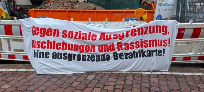 Transparent, das an einer Baustellenabsperrung angebracht ist. In roten Buchstaben steht darauf "Gegen soziale Ausgrenzung, Abschiebungen nd Rassismus!", darunter in schwarzen Buchstaben "Keine ausgrenzende Bezahlkarte!"