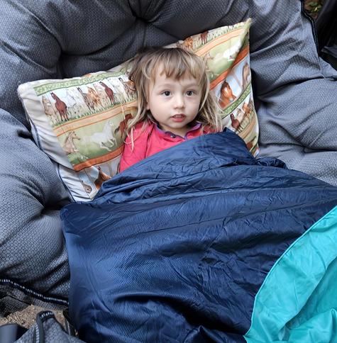 A young child with blonde hair and a red shirt is nestled inside a blue sleeping bag with a teal interior, in a large, gray, padded chair with a horse-themed pillow behind them.