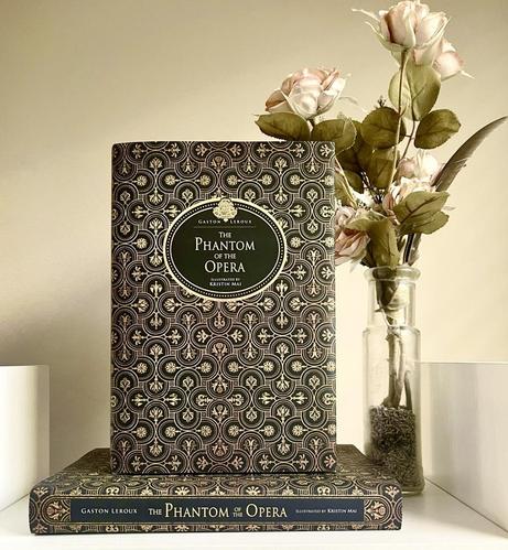 The Phantom of the Opera by Gaston Leroux and illustrated by Kristin Mai. The cover has an intricate floral gold and black pattern and there is a small golden chandelier over the title. There is a small vase of fake roses next to it.