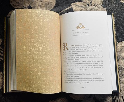 Book page spread: on the left page is a decorative chapter divider with an intricate golden design. On the right page is the start of the chapter. The chapter header has a small, gold design of a lyre with wings.