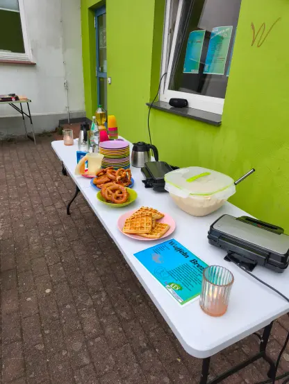 Vor der beingrünen Fassade ist ein Tisch aufgestellt. Darauf stehen Getränke, Waffeln, Brezeln und Waffeleisen