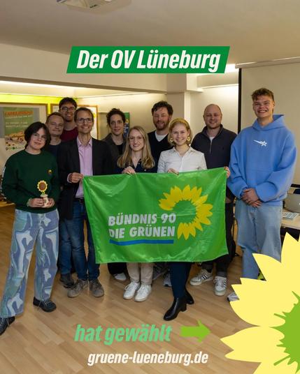 Eine Gruppe von zehn Personen steht in einem Raum auf einem hellen Holzboden. In der Mitte halten zwei Menschen ein großes grünes Banner mit der Aufschrift „BÜNDNIS 90/DIE GRÜNEN“ und einer gelben Sonnenblume. Darüber steht in grüner Schrift: „Der OV Lüneburg“. Unten rechts im Bild ist eine große Sonnenblume als grafisches Element. Daneben steht: „hat gewählt“ mit einem Pfeil sowie die Website „gruene-lueneburg.de“.