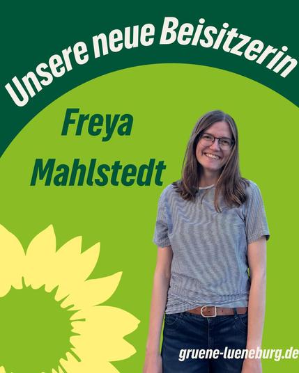 Eine Frau mit langen Haaren, Brille und gestreiftem T-Shirt lächelt in die Kamera. Sie trägt eine Jeans und steht vor einem grünen Hintergrund mit der Grafik einer gelben Sonnenblume. Oben im Bild steht in geschwungener weißer Schrift: „Unsere neue Beisitzerin“. Darunter in dunkelgrün: „Freya Mahlstedt“. Unten rechts: „gruene-lueneburg.de“.