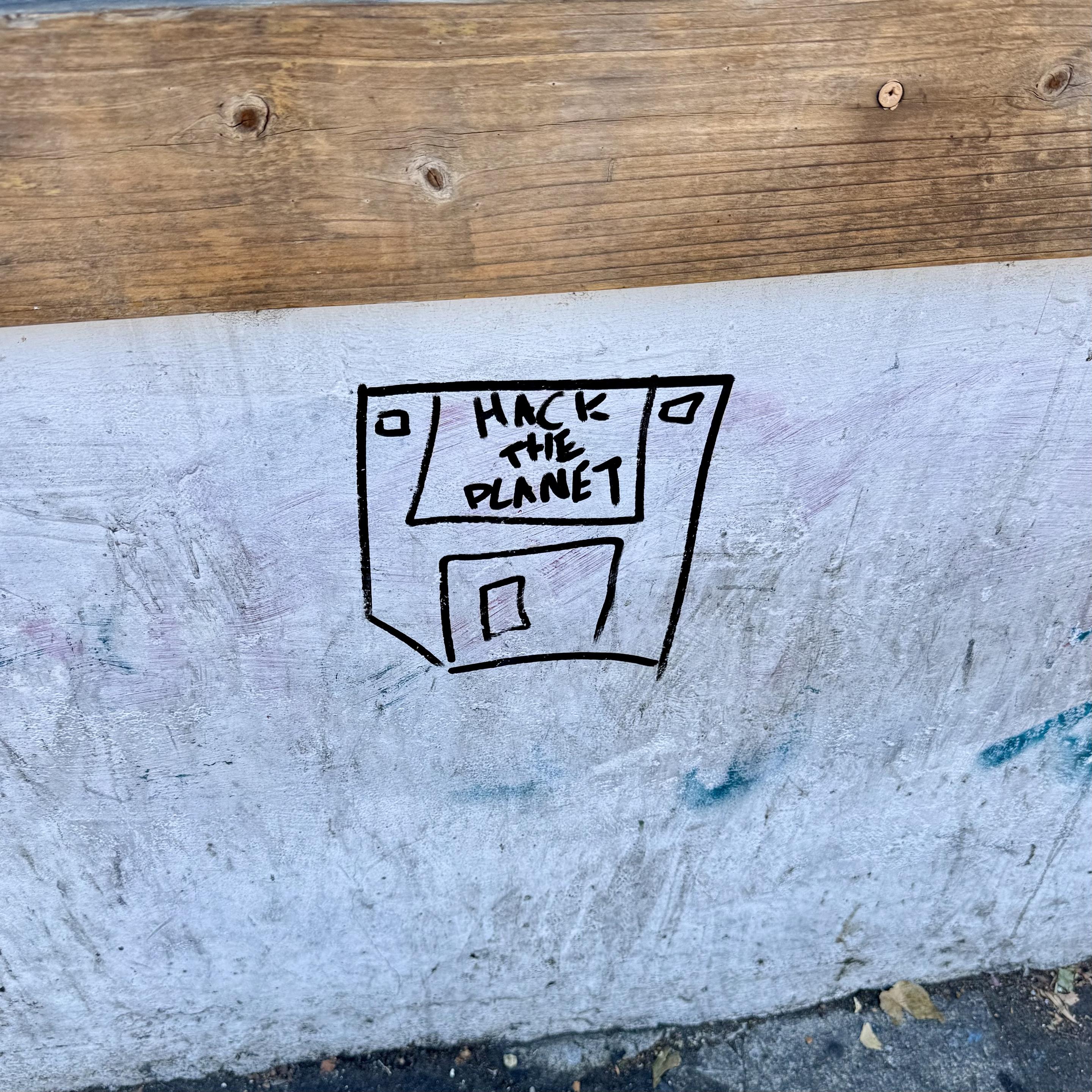 Graffiti of a floppy disk with text that says "Hack the planet". The graffiti is in black marker or paint on a white wall.