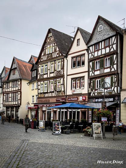 Straßenszene mit Fachwerkhäusern, Restaurants, Blumenkübeln und Passanten auf Kopfsteinpflaster.