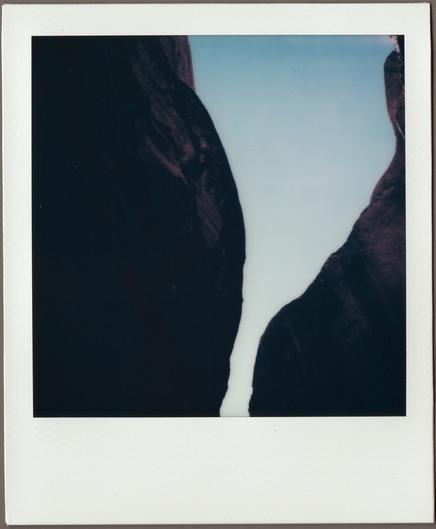 Polaroid looking up through dark canyon walls with a slice of pale blue sky showing through.