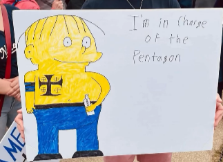 Pete Hegseth drawn as Ralph Wiggum from The Simpsons, complete with white supremacist tattoos and a bottle of something alcoholic. In intentionally crude letters, the sign reads "I'm in charge of the Pentagon."