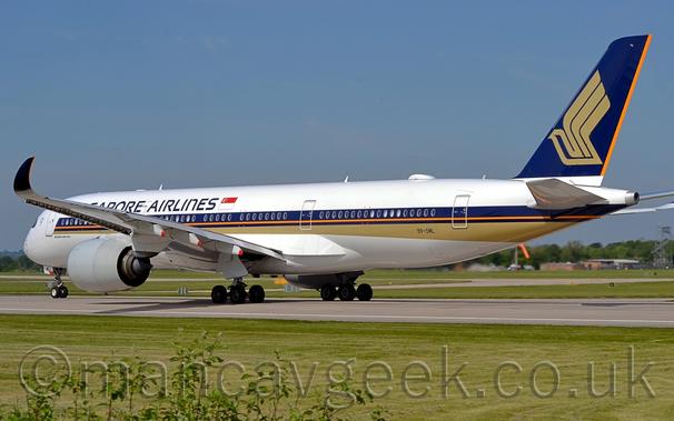 Rear side view of a twin engined jet airliner moving from right to left along a dark grey runway.
The plane is mostly white, with purple "Singapore Airlines" titles on the upper forward fuselage, and a purple and gold stripe (with a single orange pinstripe) running along the body, covering the passenger cabin windows.
The black registration "9V-SML" is on the lower rear fuselage, below the rearmost cabin windows.
The tail is purple, with a stylised golden bird with outstretched wings, and an orange stripe at the rear.
The up-turned wingtips are also purple.
 Green grass fills the foreground, lining the side of the runway, with more grass visible under the plane, on the far side of the runway.
Trees mark the airfield perimeter in the distance, under blue skies.