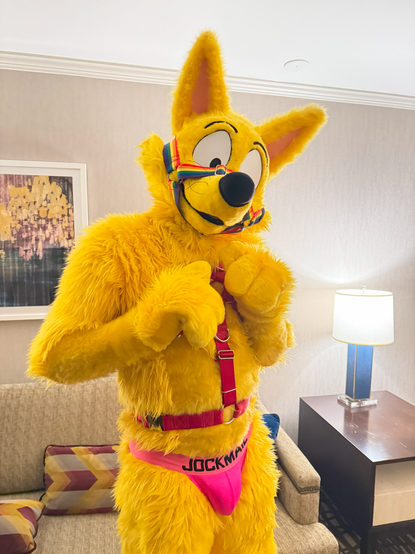 Zorori, a tall yellow fox fursuit, wearing a colorful muzzle, a harness, and a pink jockstrap. He's standing in a hotel room, looking at the camera with his head tilted.