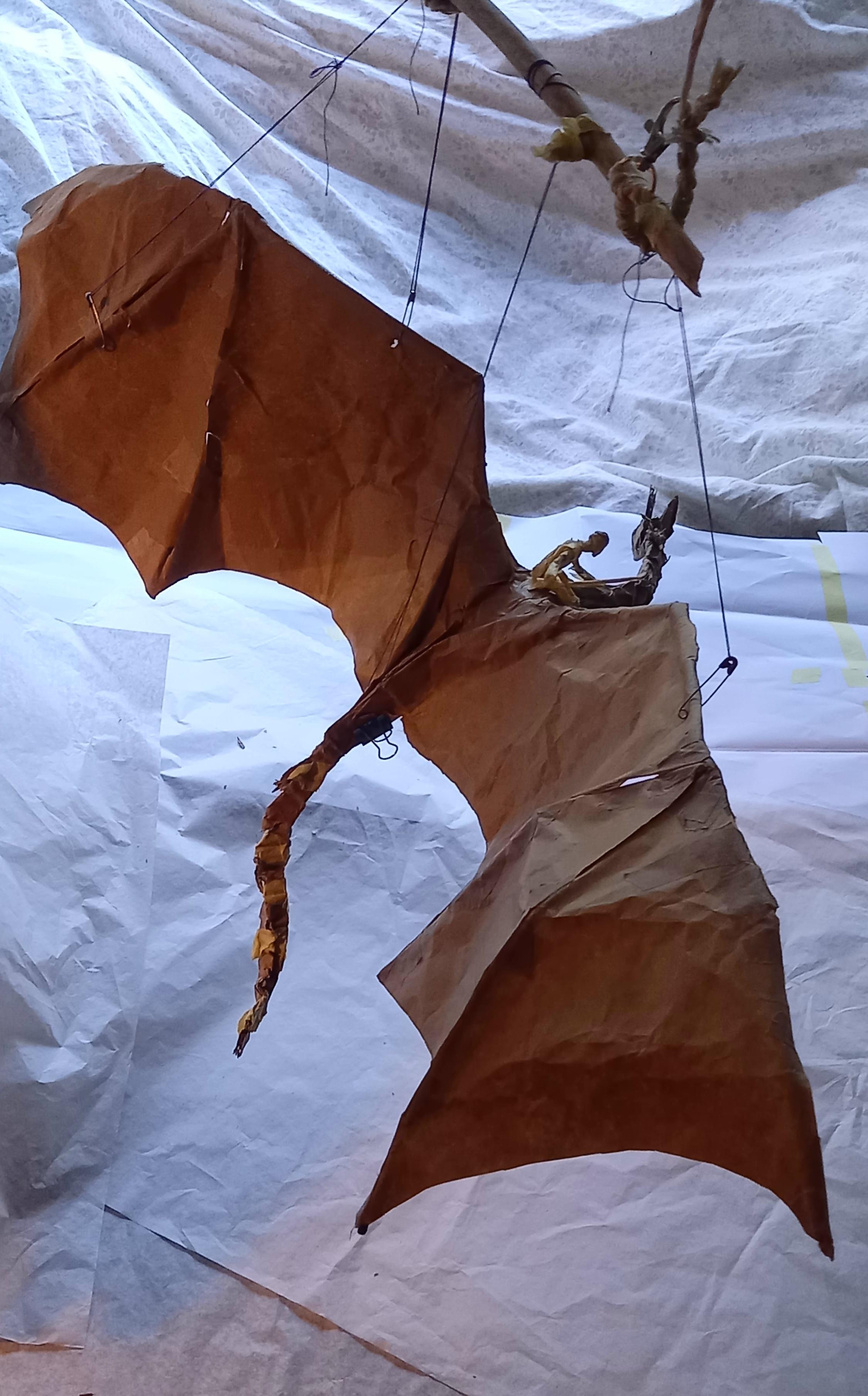 Photo of a crude paper dragon sculpture hanging from a twig in strings fastened with safety pins. White paper in the background.
