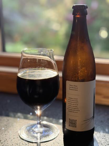 A tall brown beer bottle with a sparse white label sits bathed in sunlight. A snifter glass of the dark stout is to the left.