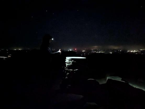 Photo of Mrs Spaceways at the top of Bennachie. Their headtorch is lighting up the stone view indicator. Stars above and distant lights of houses beyond.