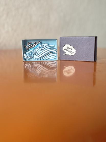 An open matchbox with a paper boat and a stormy sea on an orange surface. On the right is the outer packaging of the matchbox with a tiny speech bubble saying: happy birthday!