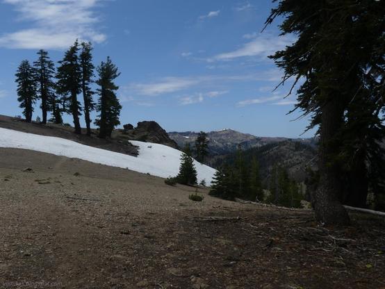 The one bit of snow on trail almost to the junction with the South Yolla Bolly Trail.
Rockyu soil of a ridge with a trail extending outward. A finger of snow crosses the trail. Trees stand in a group on the far side and off to the right on this side. Distant peaks are visible between.
