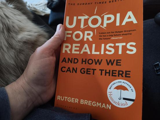 My left hand holding the book Utopia for Realists by Rutger Bregman. I'm sitting on the couch and Mia, an ancient white and tortoiseshell ragdoll cross cat, is sleeping on my outstretched legs.