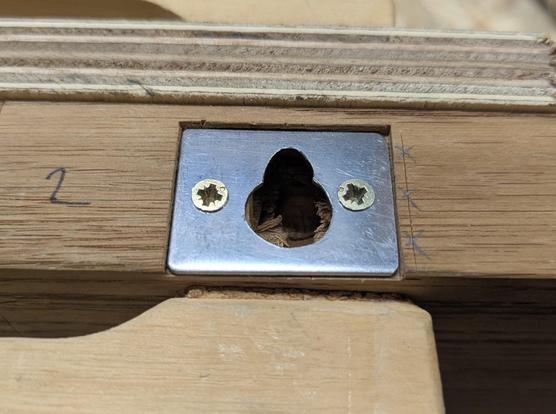 A metal hanger plate, made from a small rectangular piece of thick sheet metal. Two smaller holes at the outside affix it with screws to a shelf board. In the middle of the plate is a large hole, followed by a smaller hole above, in which the head of a screw can rest and carry the shelf. With the small screws at the side and the big honey in the middle it looks like a face with a big nose and a big open mouth.