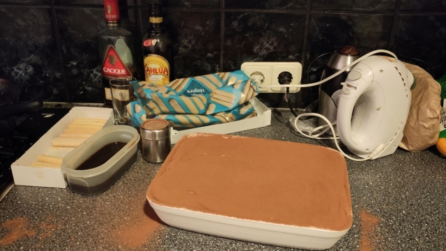 Photo of a bowl of tiramisu, with a bottle of rum and Kahlua, biscuits and coffee around it. Cocoa powder isn't only on the tiramisu, but also spilled around the bowl.
