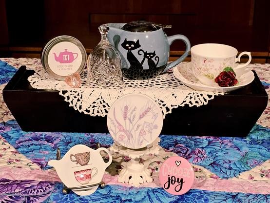 Tea tableau upon a handmade purple and teal quilt, with white and pink ceramic pieces, a crocheted lace doily, vintage china, a porcelain rose, a clear crystal bell, a blue Cattitude teapot, and Rose Peony green tea blended with rose petals and peonies.