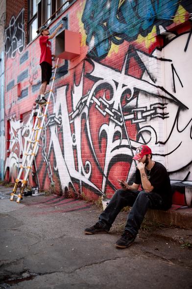 Two artists working on a new graffiti wall, one is on a ladder currently working with a paint roller, the second one is on a break sitting back to the wall, looking to his phone while smoking a cigarette.

The wall is red with white lettering and black outlines. Amusingly both artists are also wearing red and black.