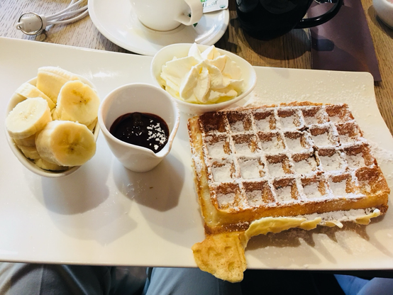 A meal that consists of sliced banana, chocolate sauce, Belgian waffle with powdered sugar on top and butter. Served at a small cafe in Ypres, Belgium.