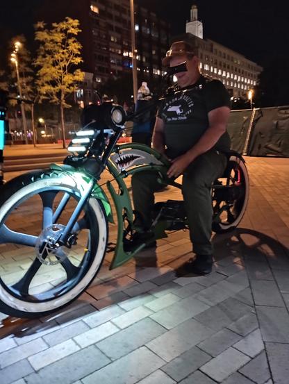 Guy sitting on a low rider bicycle.  The headlight is shining off the front tire. Night time, high rise buildings in the background.