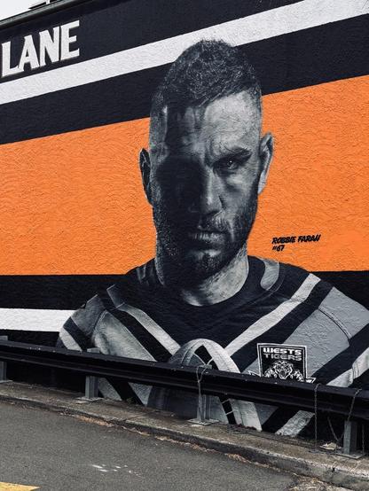 Street art mural on a wall by a laneway of Robbie Farah #67 for Wests Tigers. He looks directly at the viewer with an expression of “I’ve got the ball and there is no way you’re getting it off me”. He is in dramatic black and white, with the background of the wall in bright orange.