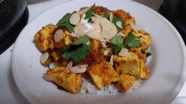Assiette blanche avec au centre un plat indien, du tofu tikka masala Les cubes de tofu colorés par des épices reposent sur du riz et sont garnis de coriandre, d'amandes effilées, de yogourt et de poivre.
