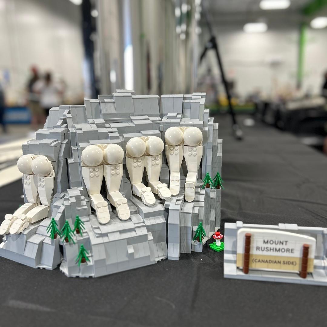 as Bild zeigt eine Lego-Darstellung von Mount Rushmore, mit einer humorvollen Variation. Im Vordergrund befindet sich eine grüne, steinige Landschaft mit mehreren Lego-Figuren, die als Hüften und Beine dargestellt sind, anstatt der tatsächlichen Porträts der Präsidenten. Diese Figuren sind in einer Reihe angeordnet und sitzen auf einem felsigen Hügel. Kleine grüne Bäume sind um die Figur gruppiert. Vor der Darstellung steht ein Schild mit der Aufschrift "MOUNT RUSHMORE (CANADIAN SIDE)". Die Hintergrundumgebung ist einigermaßen unscharf, mit einem professionellen Umfeld, das auf eine Ausstellung oder einen Workshop hinweist.