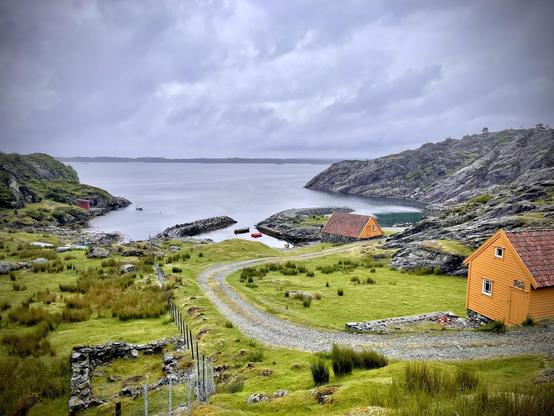 Overcast sky above a rugged coastal landscape with a winding gravel road leading towards two orange wooden houses near the shore. Rocky cliffs border a calm bay, with a small red boat floating near a jetty, enhancing the serene rural setting. Green grass and sparse trees are visible around the houses, depicting a tranquil, isolated environment.