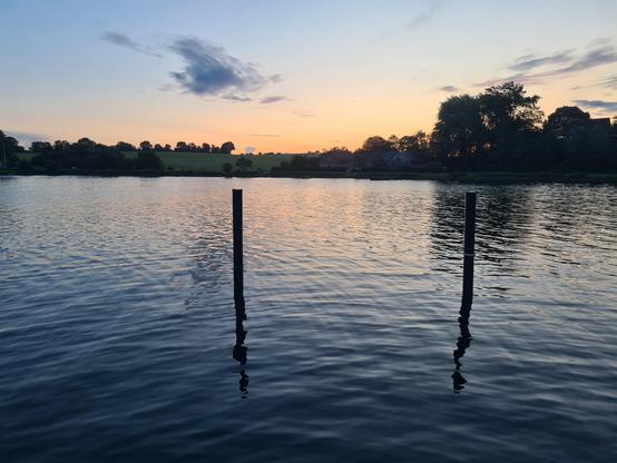 Blick auf einen Sonnenaufgang hinter grünen Hügeln, davor eine Wasserfläche mit zwei Pfosten die herausragen