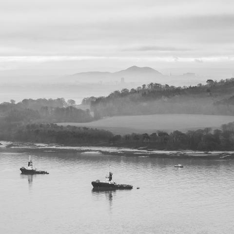 A black and white photo of a view towards Edinburgh from atop the Forth Bridge. A couple of tugboats are anchored in the still water of the foreground, the wooded shoreline behind them. In the distance, riding through the mist, Arthur’s Seat indicates the location of the otherwise largely hidden city of Edinburgh.
It’s still there, though, right? RIGHT?
