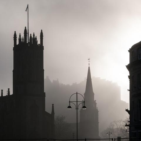 A black and white photo of Edinburgh on a foggy day. The silhouetted spiky towers and spires of a couple of churches claim the foreground, with layers of trees or other rooftops behind gradually fading into a hazy mist. Behind them all, the enormous mass of the castle looms high above.
If it weren’t for the street lights, this could have been taken hundreds of years ago. It’s weirdly quiet… have we travelled back in time??
