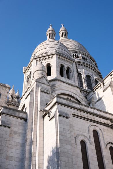 Lower angle pic on a part of the Sacrée Cœur in Paris. We can see 2 towers with domes as roof. The white bricks bring a lot of contrast with the deep blue sky. The sun shines on the building from the left